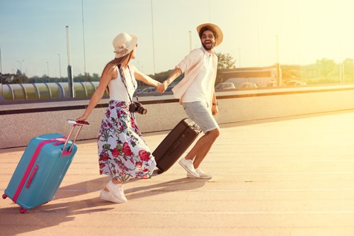 Woman in flower skirt and white hat dragging a blue suitcase holds the hand of a man in shorts, white shirt and hat dragging a black suitcase
