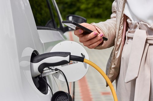 Woman holds mobile phone, standing by a car plugged in to an EV charging point