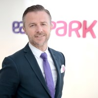 Businessman wearing a blue suit jacket, white shirt and purple tie standing in front of the EasyPark logo