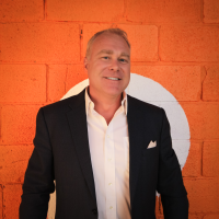 Business man in a black blazer and open neck white shirt stands against an orange wall