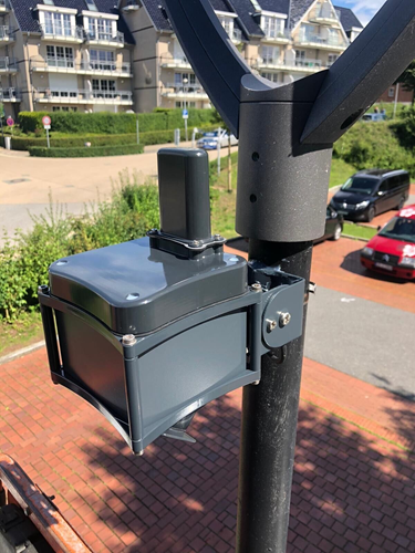 A black parking sensor attached to a lamppost in a car park, with buildings behind