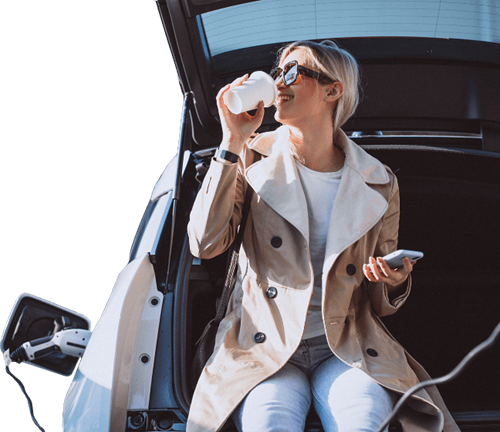 Woman sits in the trunk of the car, drinking coffee and using an app whilst her car charges