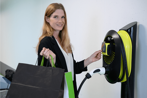Woman with shopping bags taps a payment card against an EV wall charger