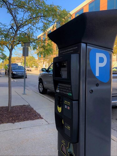 Black, solar-powered parking meter with key pad on sidewalk with trees, cars and a building in background