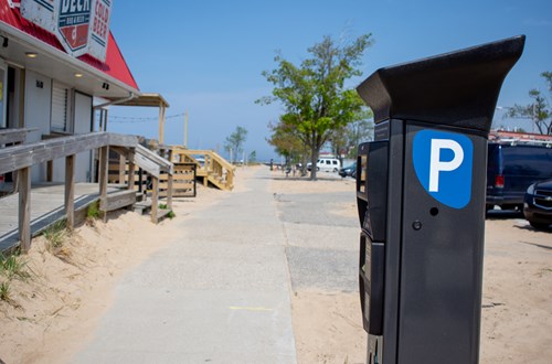 The Flowbird system charges visitors for parking whilst residents have a parking permit to access the beach for free. A parking meter in the foreground with a boardwalk along a beach and on-street parking.