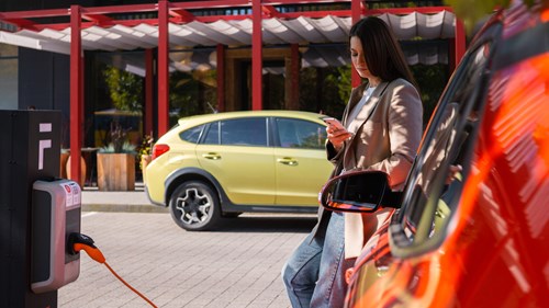 A woman leans against a red electric vehicle as it charges, yellow car drives past in background