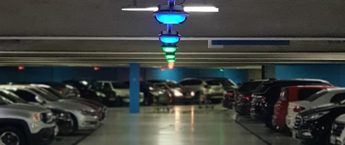 Interior of a parking garage with white floor and blue columns. A line of parking guidance sensors on the ceiling are illuminated blue, green and red.