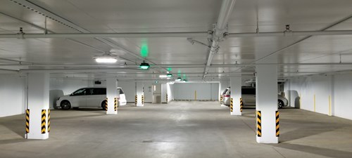 Well lit white parking garage with a silver car and white car parked in bays and green parking guidance sensors.