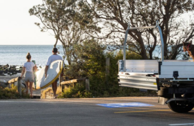 A couple leave a truck in a car park to walk down to the beach, surfboard in hand