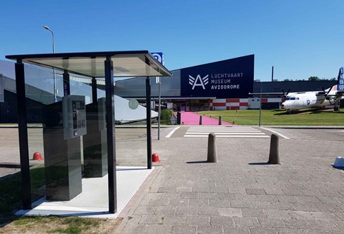Parking payment terminals in a glass shelter with an airport museum and airplane behind