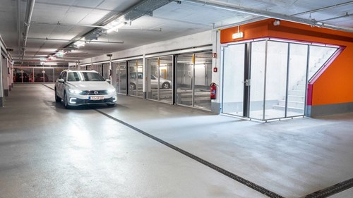 Car drivers through a grey and orange parking garage, with cars behind parking lift cages